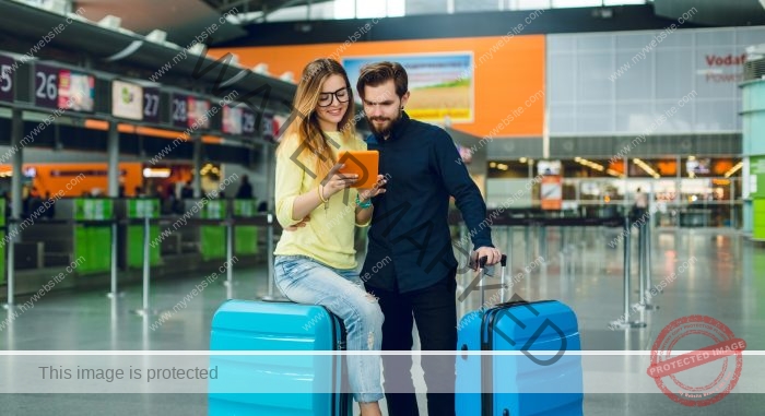 Young girl with long hair in yellow sweater, jeans is sitting on suitcase in airport. Guy with beard in black shirt with pants and suitcase is standing near. They are looking on tablet.